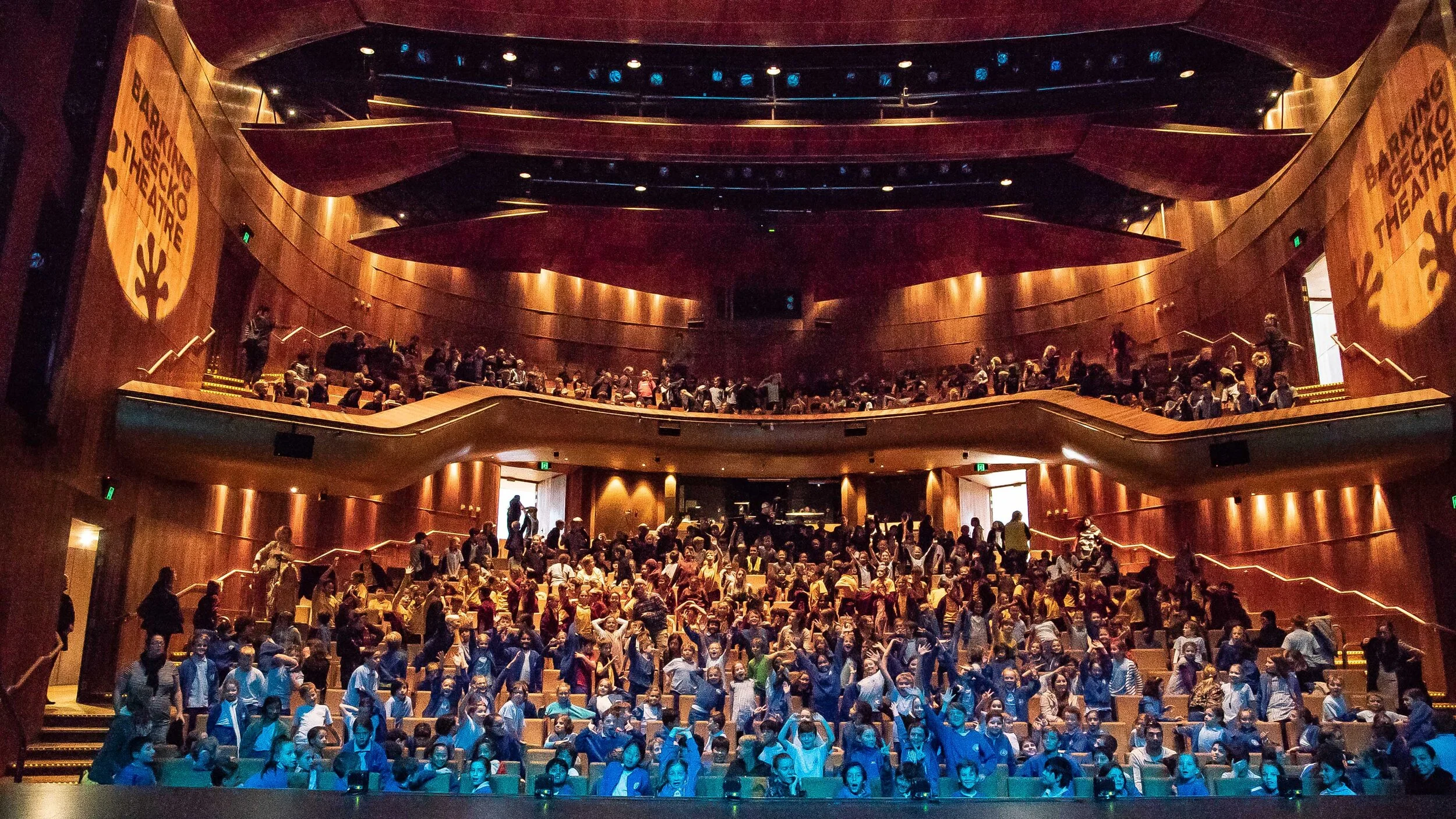 A packed theatre auditorium with school audiences in blue uniforms, warm amber lighting on the architecture and balconies