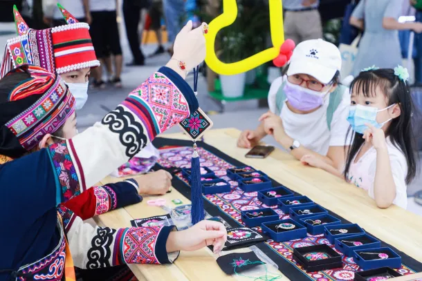 Children engaging with traditional Gansu Province textiles and cultural crafts at a community cultural event, with colorful embroidered fabrics and interactive learning