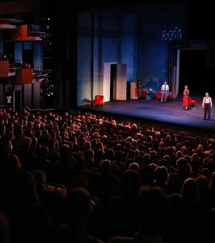 A full theatre auditorium with audiences in warm amber light, watching actors on stage in a modern production with minimalist set design