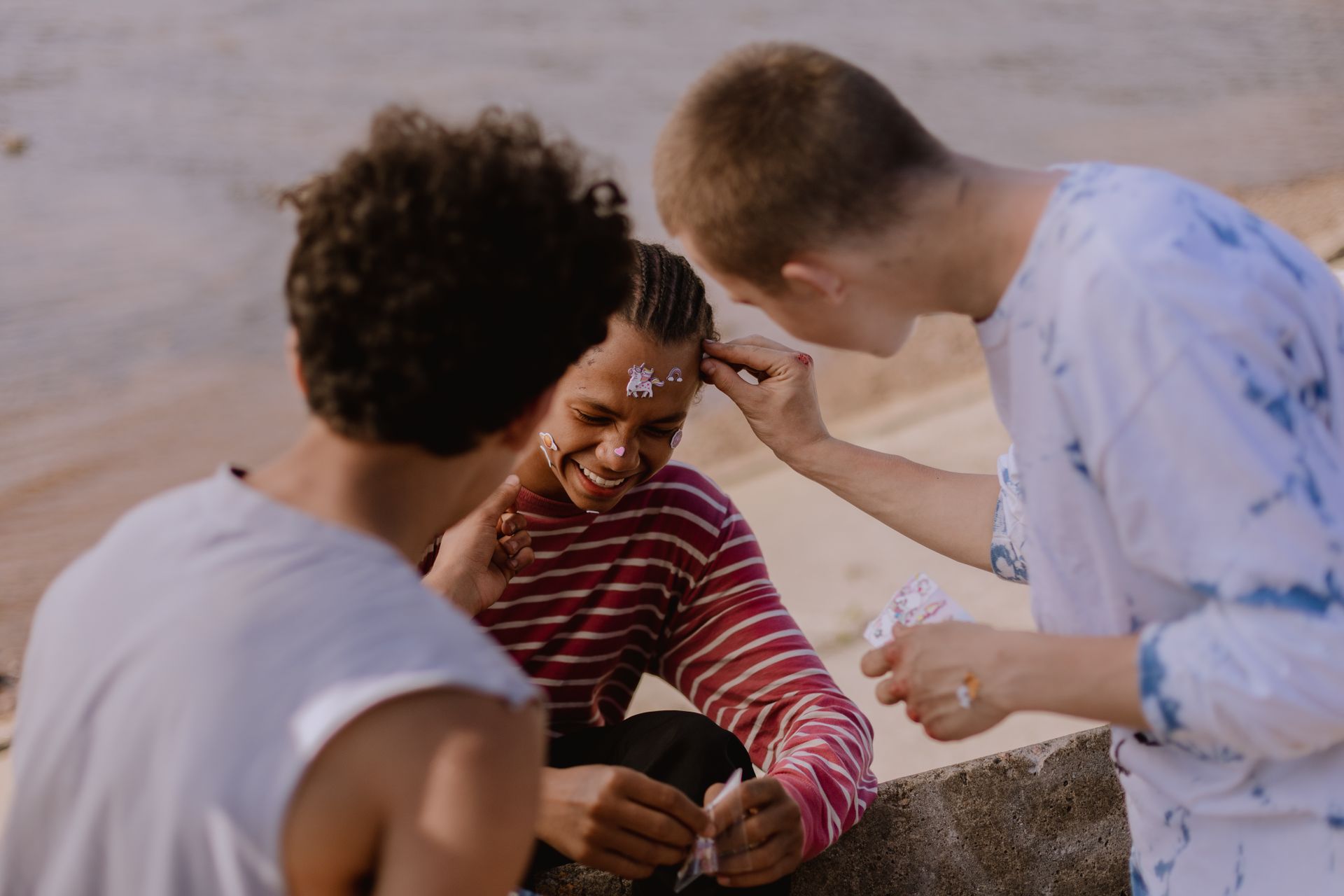 Two educators and a child in a warm moment of arts learning outdoors, with face painting and joyful engagement on a sandy beach setting