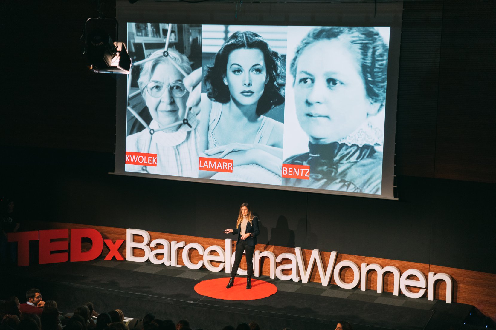 A speaker on the TEDxBarcelonaWomen stage with portraits of women innovators projected behind her