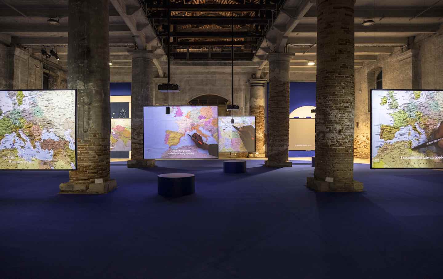 A Venice Biennale exhibition space with monumental brick columns, digital displays showing maps and cultural imagery, dramatic blue lighting and architectural grandeur